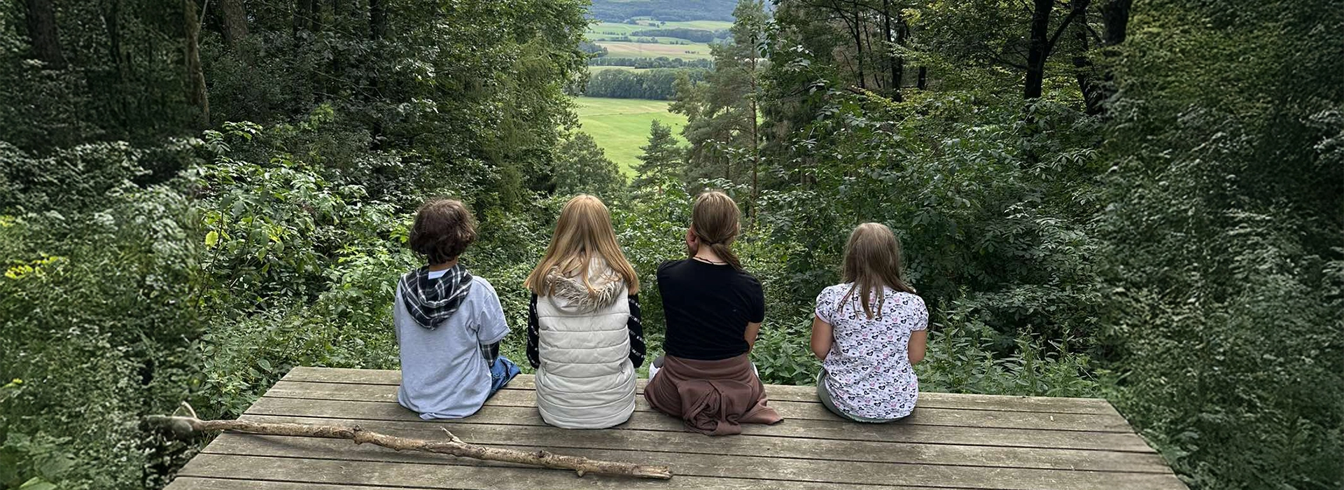 Vier Kinder sitzen im Wald auf eine Holzplattform und schauen ins Tal.