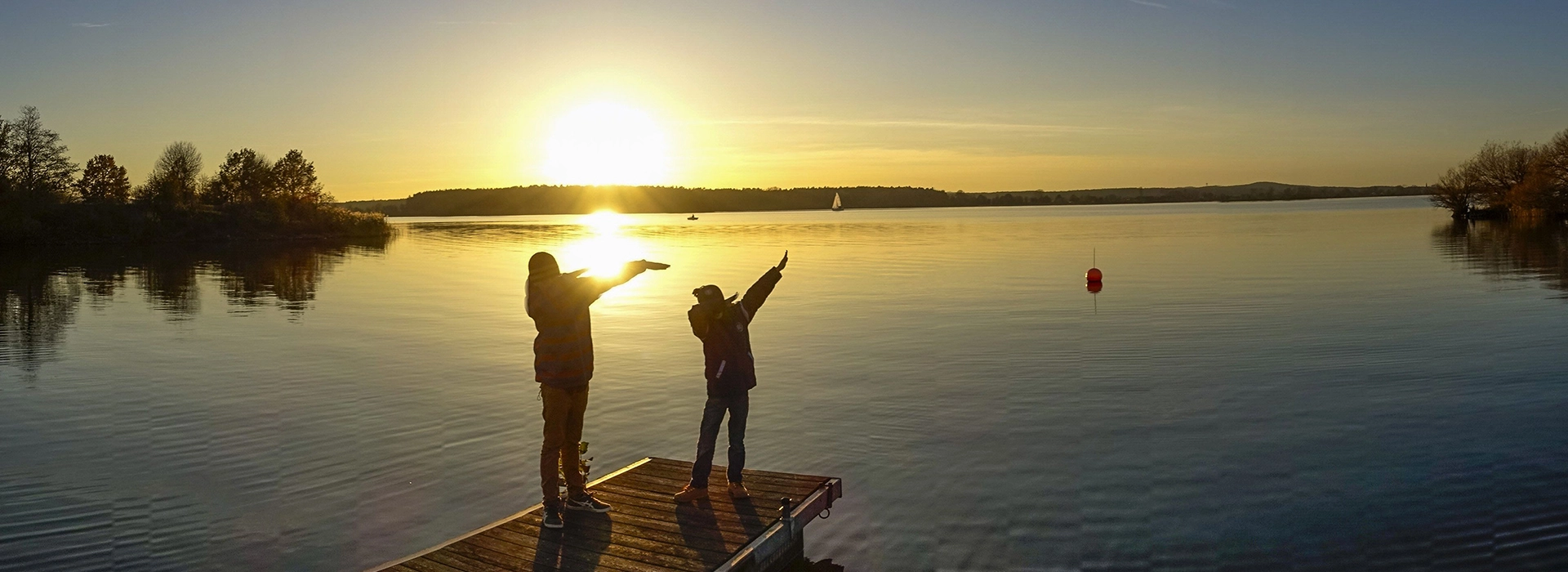 Zwei Kinder stehen bei Sonnenuntergang am Steg an einem See.