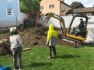 Ein Hausmeister baggert ein Loch für die neue Schaukel. Zwei Kinder schauen aufmerksam dabei zu.