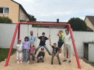 Zwei Mitarbeiter und mehrere Kinder sind bei der neuen Schaukel versammelt.