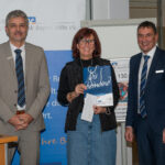 Zwei Herren von der VR Bank stehen neben einer Frau, die einen Spendenumschlag und eine Geschenktüte zeigt.