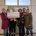 Sechs Personen stehen in einem Gang mit einem Scheck vor sich zu einer Spende