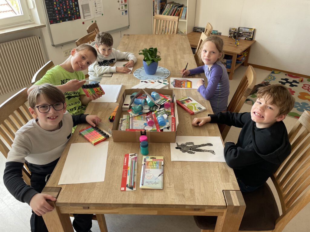Fünf Kinder unterschiedlichen Geschlechts sitzen an einem großen Holztisch mit einer Kiste Malsachen und angefangenen gemalten Bildern vor sich