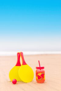 Das Bild zeigt zwei Tischtennisschläger samt Ball und einem Getränk am Strand.